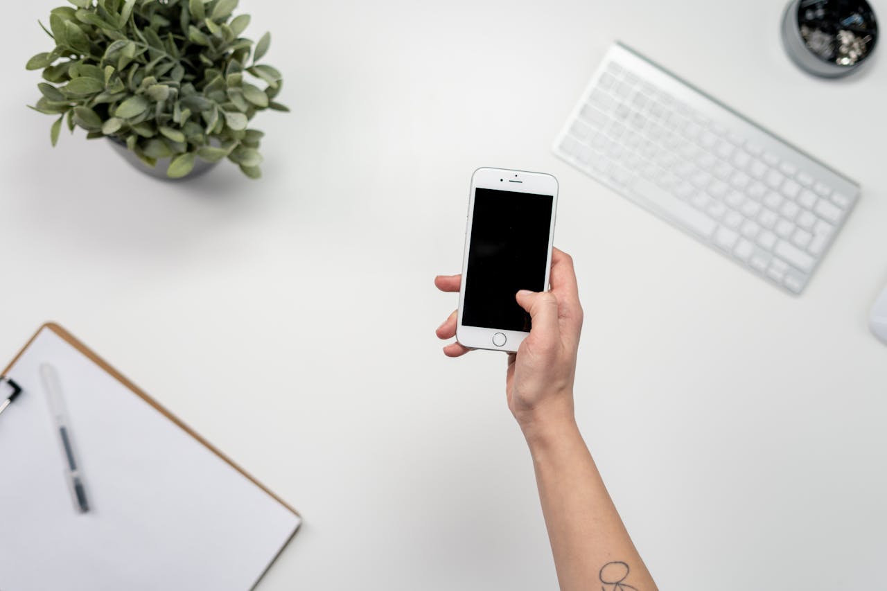 Beyaz masada iPhone tutan el, yanında klavye ve not defteri, telefon kılıfı odaklı masaüstü.